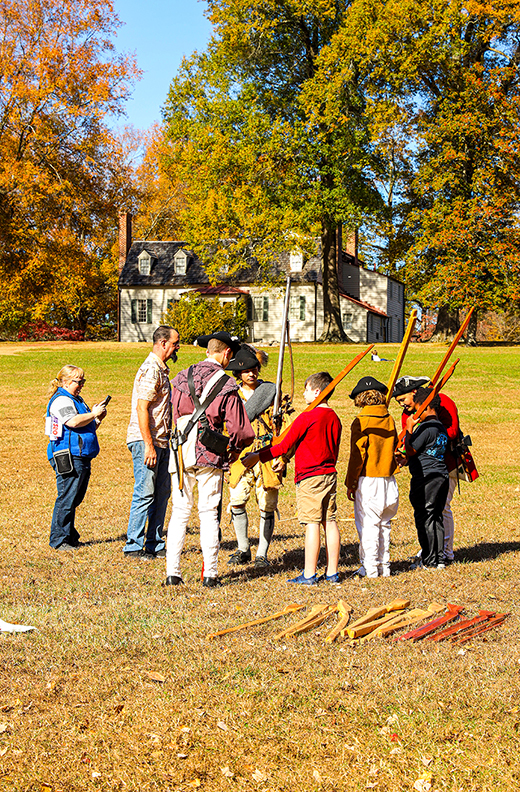 A Revolutionary War reenactor visits with several adults and children, who hold props that resemble muskets.