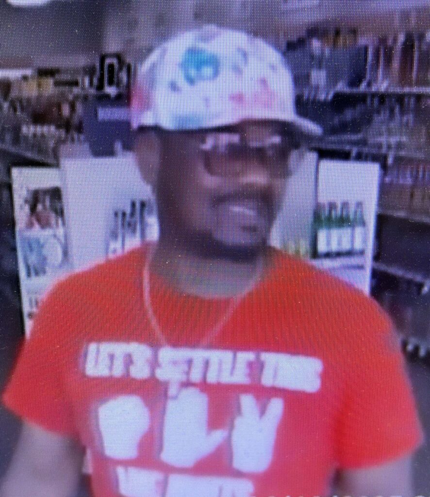 Surveillance screenshot of a person inside an ABC store. They're wearing a red shirt and white hat.