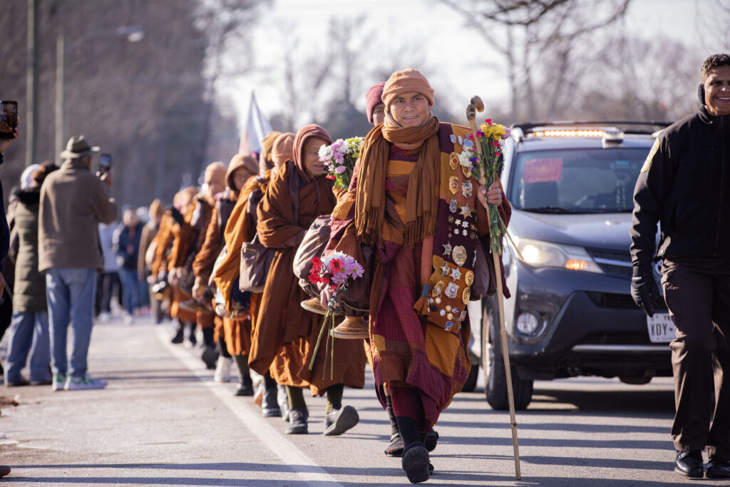 A line of 19 monks walk along a road carrying flowers.