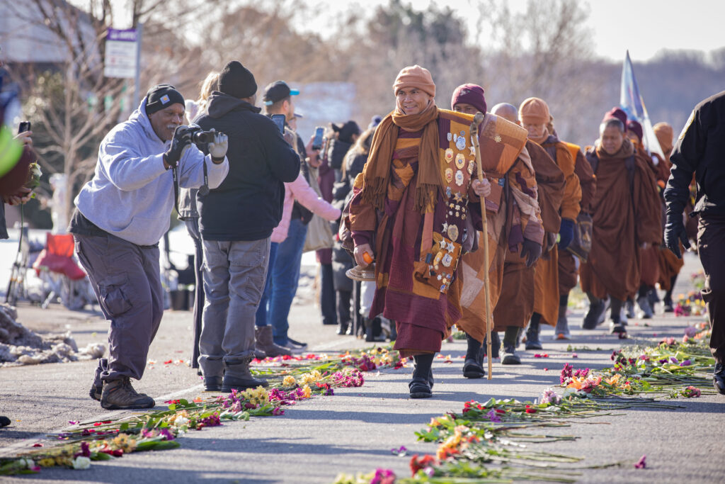 A photo of monks on their Walk for Peace are greeted by supporters and walk on a path created by flowers placed on the road.