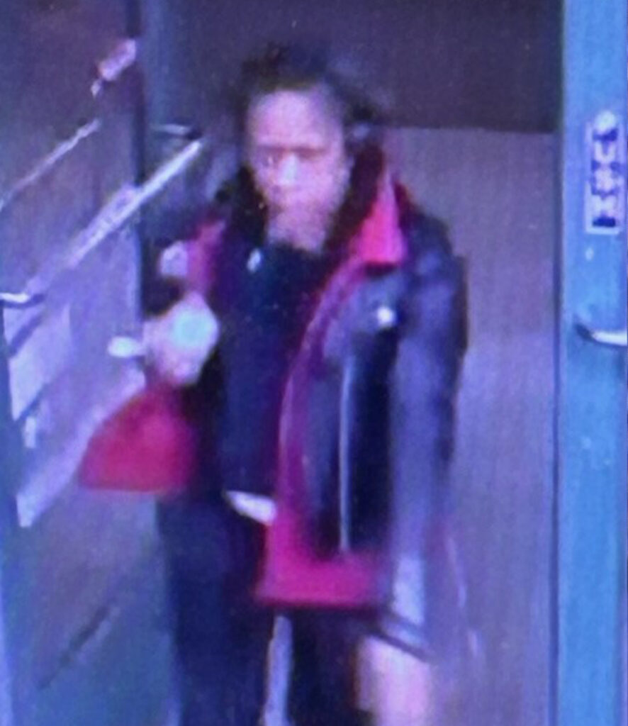 Person wearing a black leather jacket with red accents walking through a doorway indoors.

