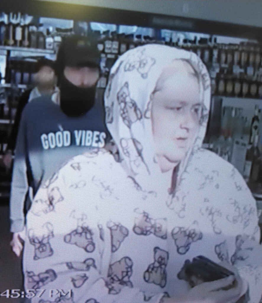 Person in a white hoodie with teddy bear prints holding a dark object inside a store with shelves of bottles behind them.
