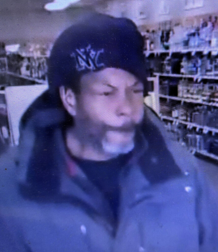Person wearing a dark jacket and black cap inside a store aisle filled with various products on shelves.