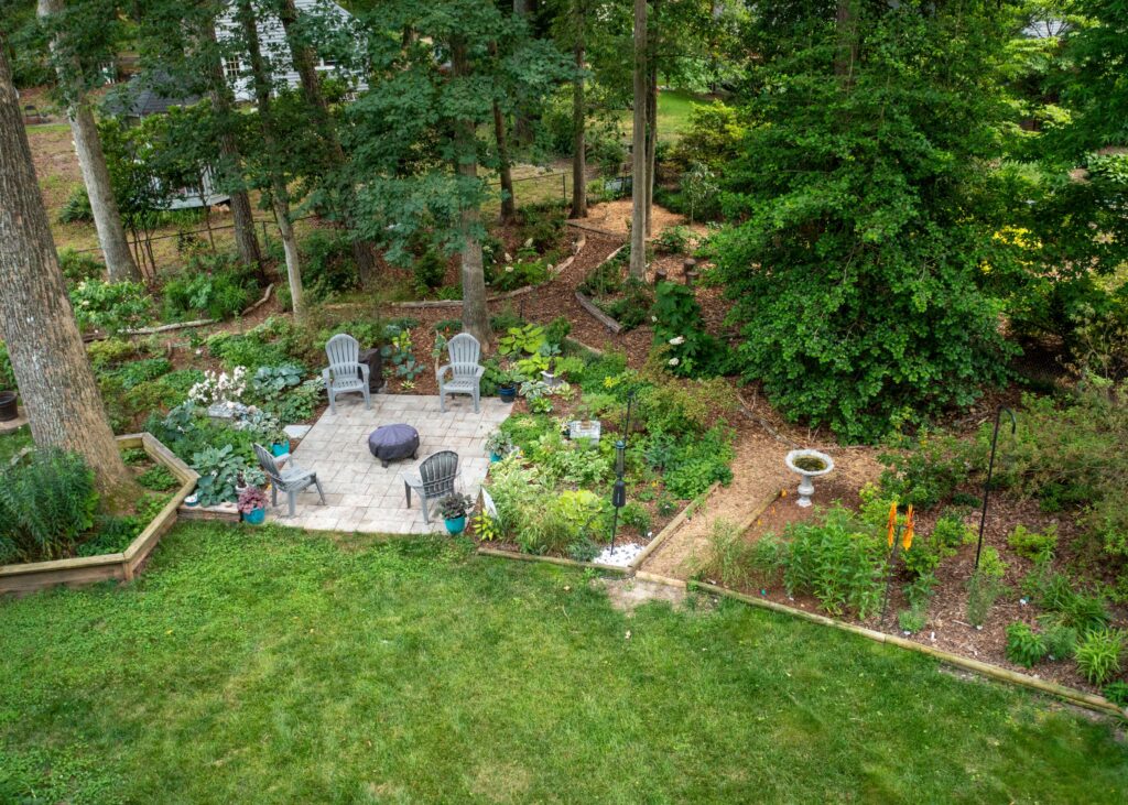 A paved patio with deck chairs surrounded by many green shrubs, pine needle paths, and trees.
