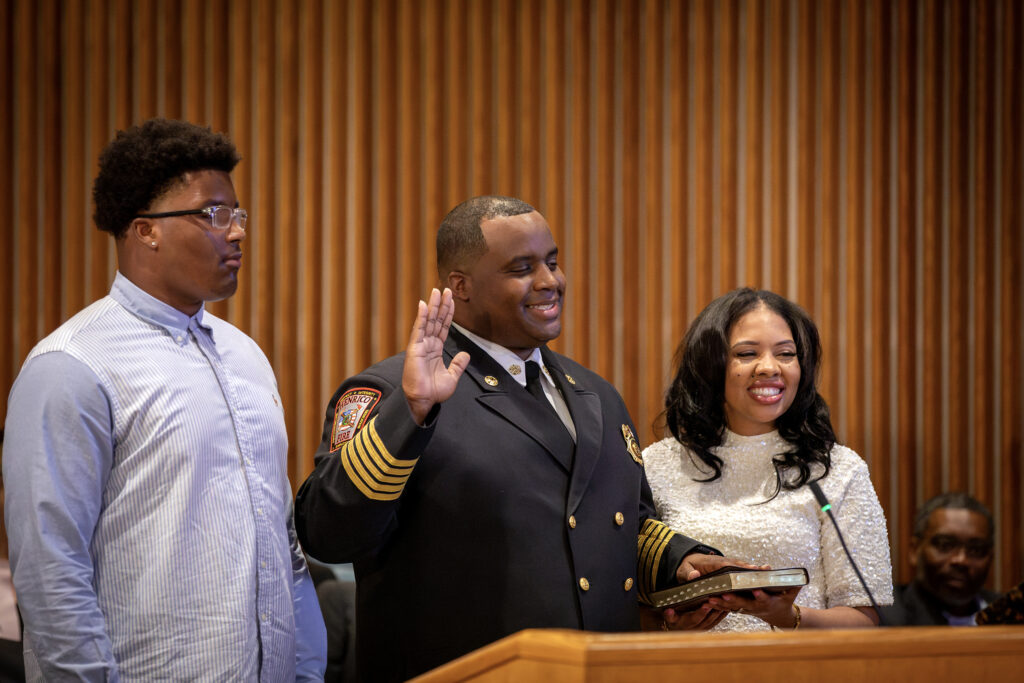 In a formal meeting room, a man raises his right hand to take the oath of office as Fire chief. His wife and son smile beside him.
