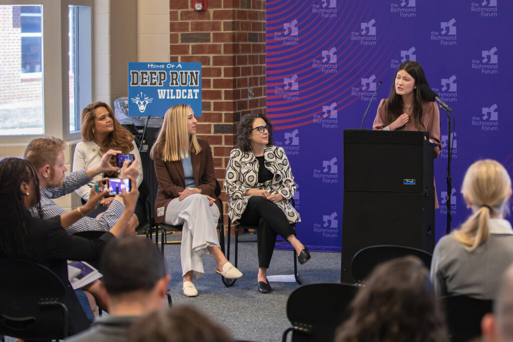 A high school student speaks at a lectern at a school with an audience of adults.