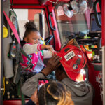 A little girl sits in a fire engine and touches a fire helmet on a man’s head.