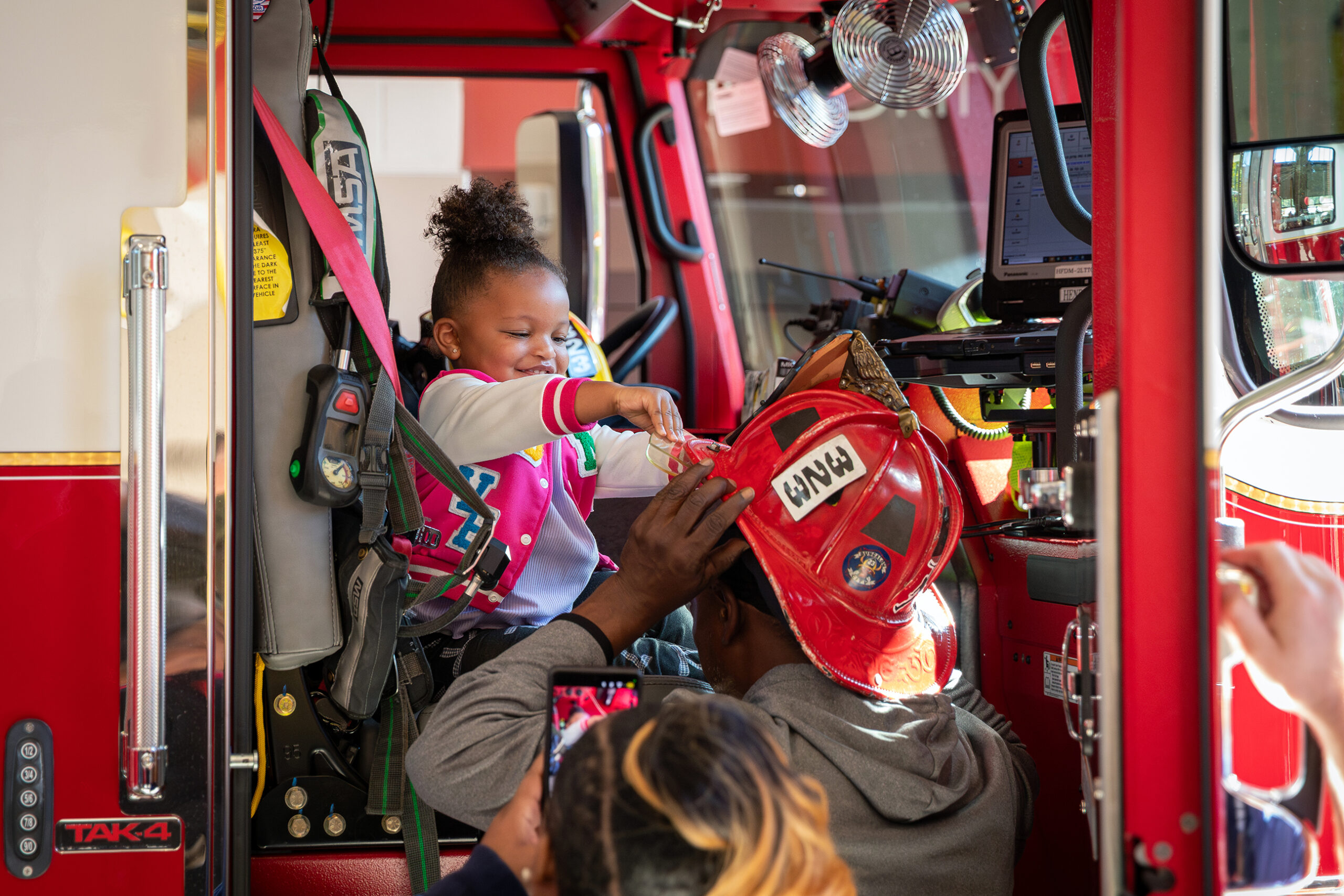 A little girl sits in a fire engine and touches a fire helmet on a man’s head.