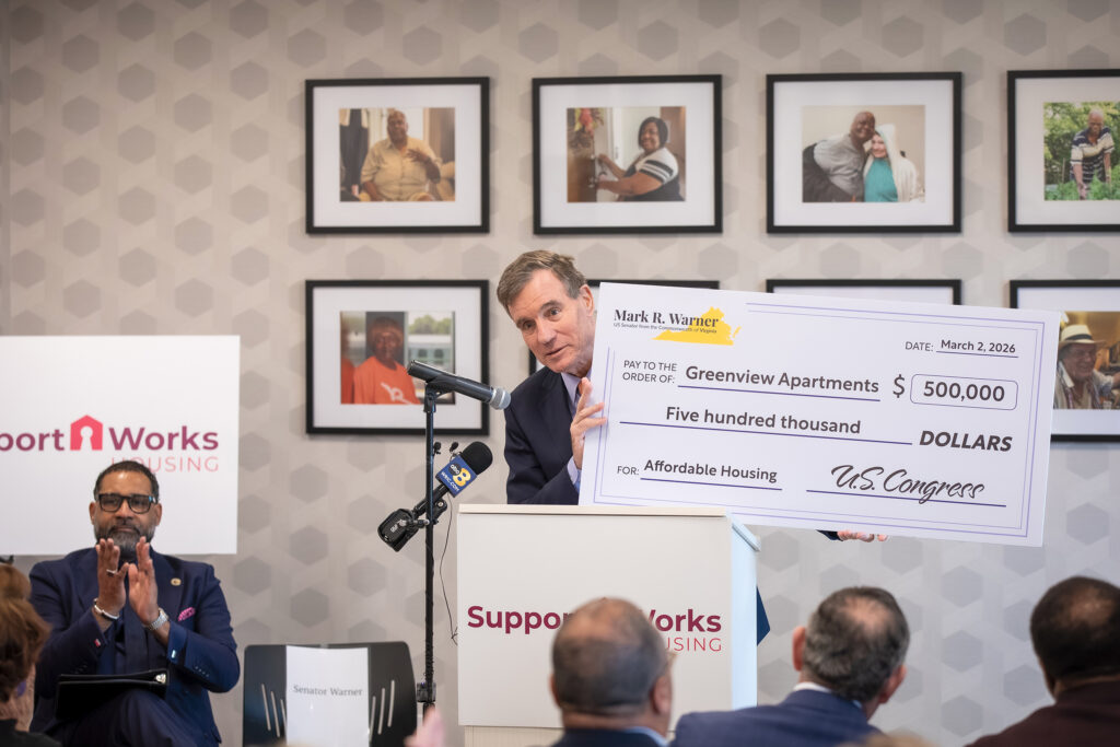 Speaking to a seated audience, Senator Mark Warner holds an oversized check for $500,000 for Greenview Apartments.