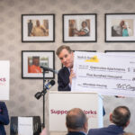 Speaking to a seated audience, Senator Mark Warner holds an oversized check for $500,000 for Greenview Apartments.