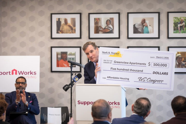 Speaking to a seated audience, Senator Mark Warner holds an oversized check for $500,000 for Greenview Apartments.