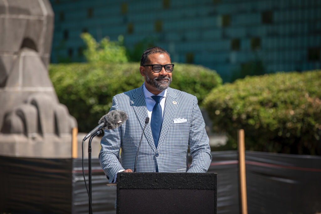 An official smiles while speaking at a lectern at an outdoor event.