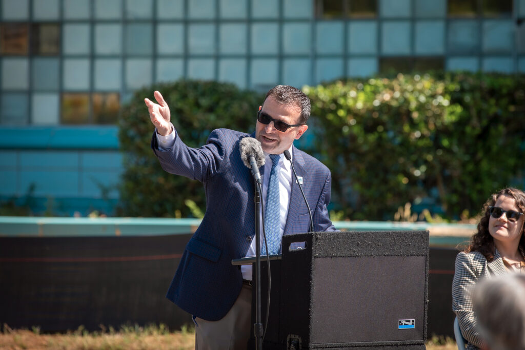 An official gestures while speaking at a lectern at an outdoor event.