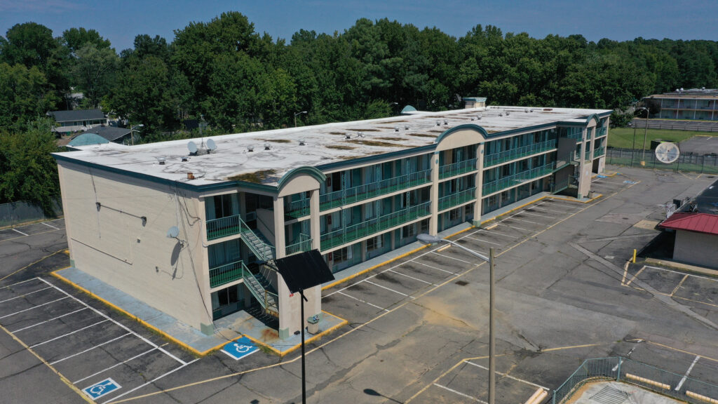 Aerial view of a three-story, beige motel with glass balconies. The building is surrounded by an empty parking lot with marked spaces, including accessible parking. A wooded area can be seen in the background.