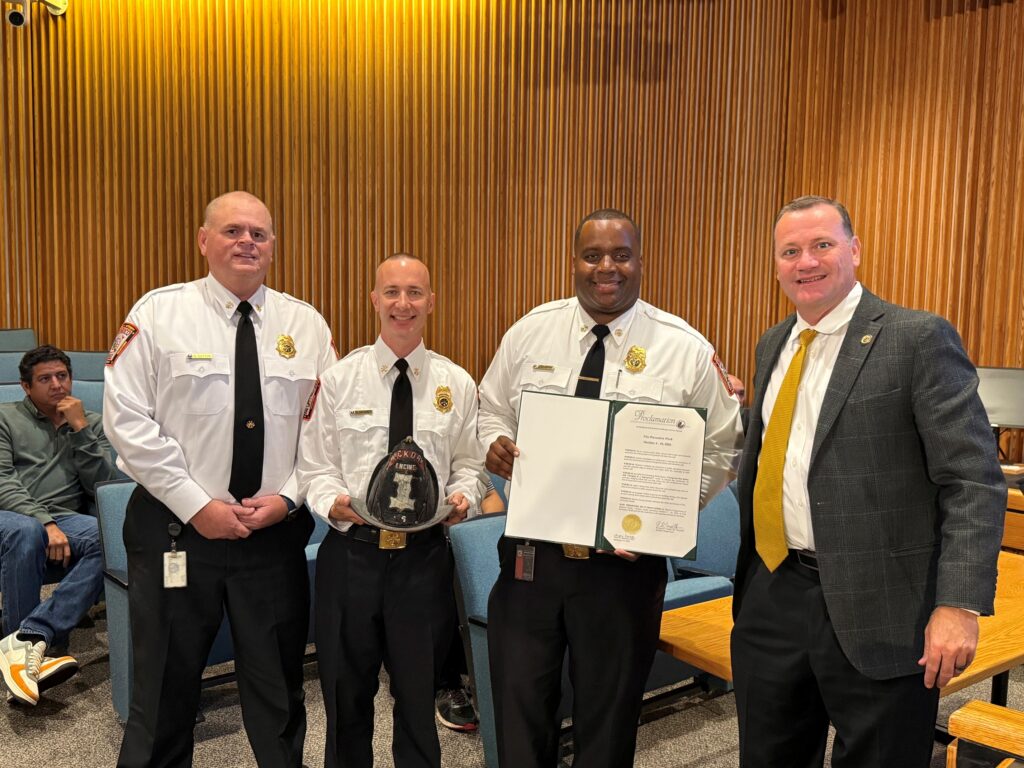 Three fire officials and an elected official pose with one hold a proclamation for fire prevention week.