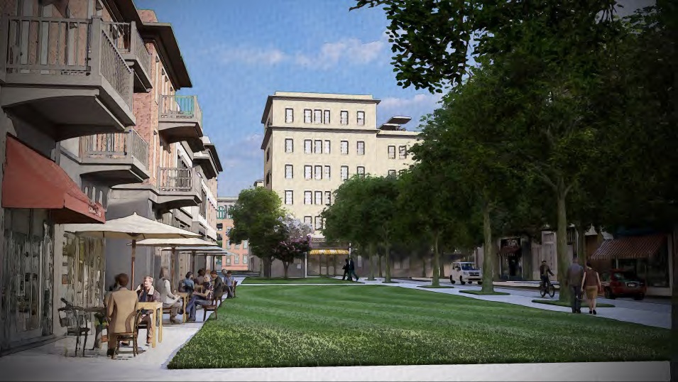 A street scene with a green park area lined with trees. People are sitting at outdoor tables under umbrellas on the left side near shops and cafes. A multi-story building is visible in the background, and a few cars are parked along the street. The sky is clear and blue.