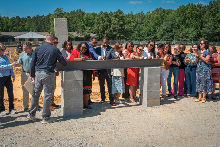 A group of people stand together holding a metal beam at a construction site. Some individuals sign the beam. Trees and a partially built structure are visible in the background under a clear blue sky.
