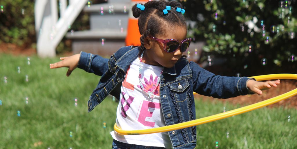 Young girl will sunglasses and a jean jacket spinning a yellow hula hoop.