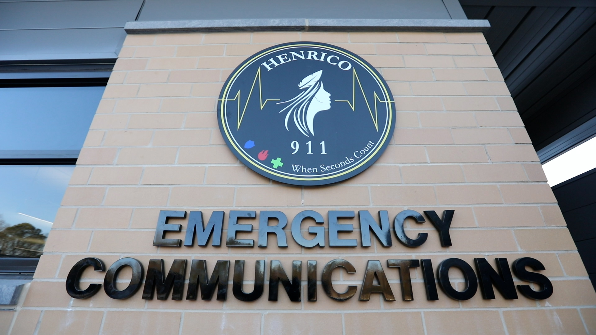 Big circle sign logo of the Henrico Department of Emergency Communications on the side of a building. Black text and mostly black and white logo with the silhouette of Pocahontas.