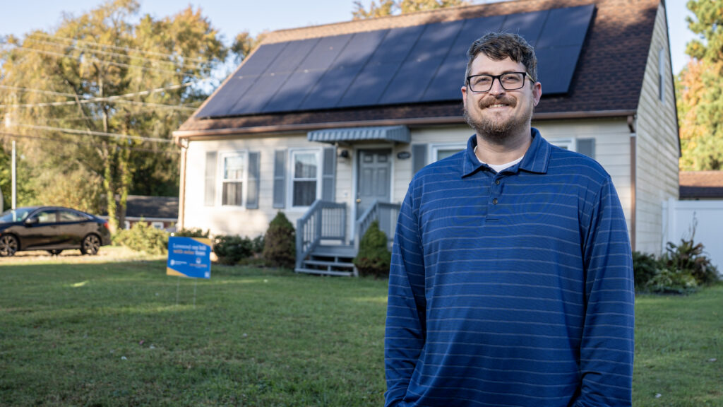 A man stands in front of a house with solar panels on the roof.