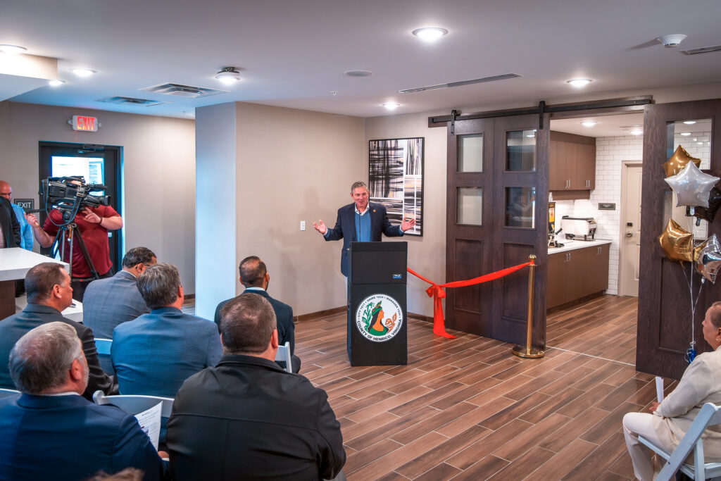 A man in a suit speaks at a lectern in front of a small seated crowd before a ribbon-cutting ceremony.