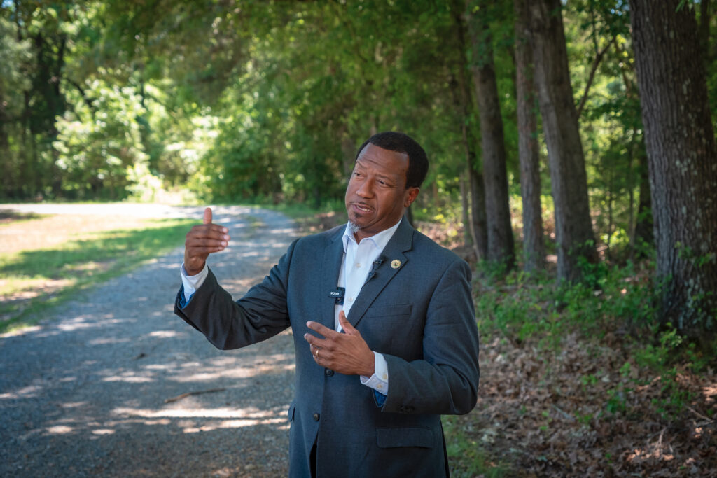 An official in a suit speaks to a reporter with woods and a gravel road in the background.
