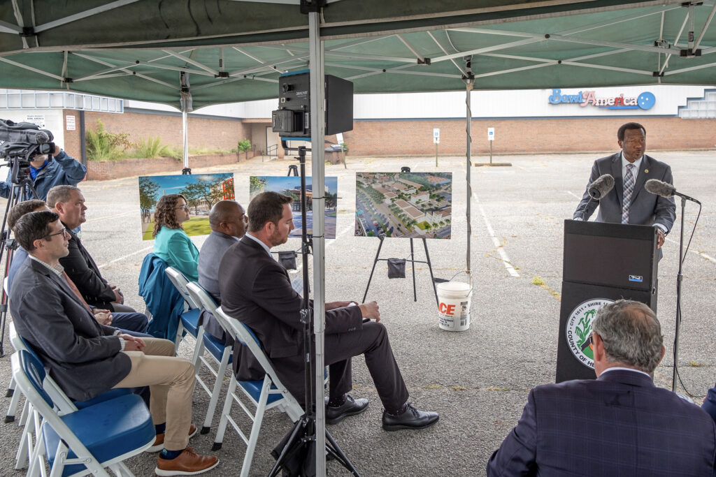 A group of people sit under a canopy listening to a speaker at a podium. The speaker stands in front of two microphones with a county seal displayed. Behind the speaker are display boards with images of a development plan. The event takes place in a parking lot with a building featuring "Bowl America" signage in the background. Chairs and a camera are set up facing the speaker.