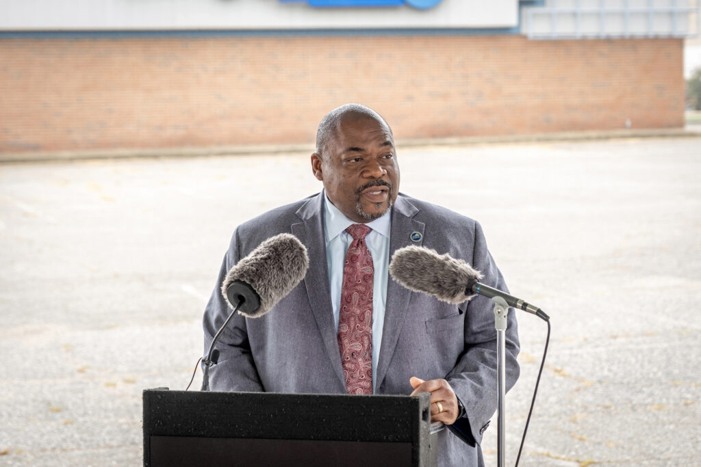 A man speaks at a lectern in front of a vacant parking lot.