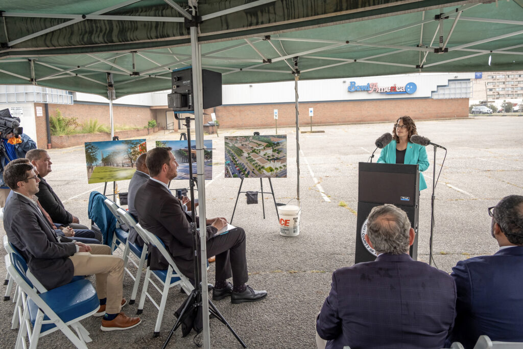 A person stands at a lectern under a green tent, speaking to a seated audience in an outdoor parking lot. Behind the speaker are images of architectural plans and a building with the sign “Bowl America."