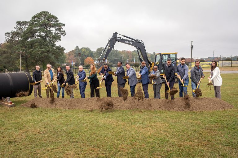 A large group of people use gold shovels to throw dirt from a pile in front of an excavator.