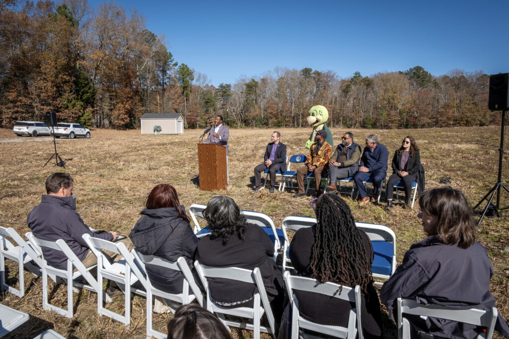 Varina Supervisor Tyrone Nelson speaks at a lectern. A small audience and a person in a turtle mascot costume are also shown in the middle of a farm. 