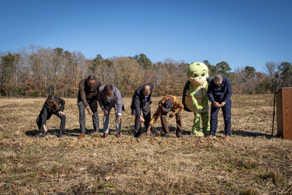 Officials scatter seeds to celebrate the start of a community farm. A person in a turtle mascot costume looks on.