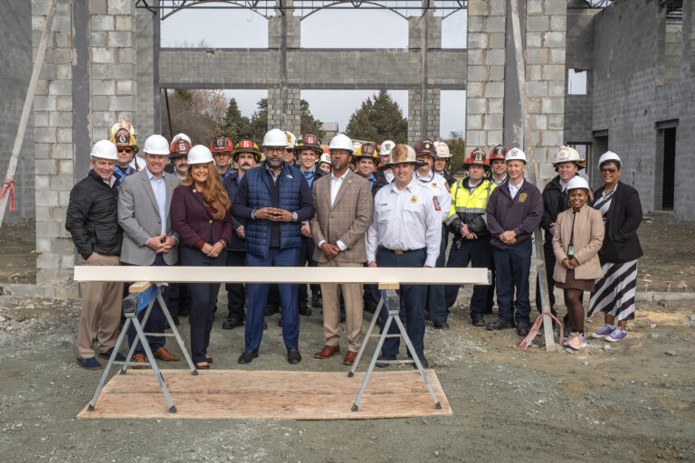 A group of more than 20 adults wear hard hats and pose for a photo at a construction site.