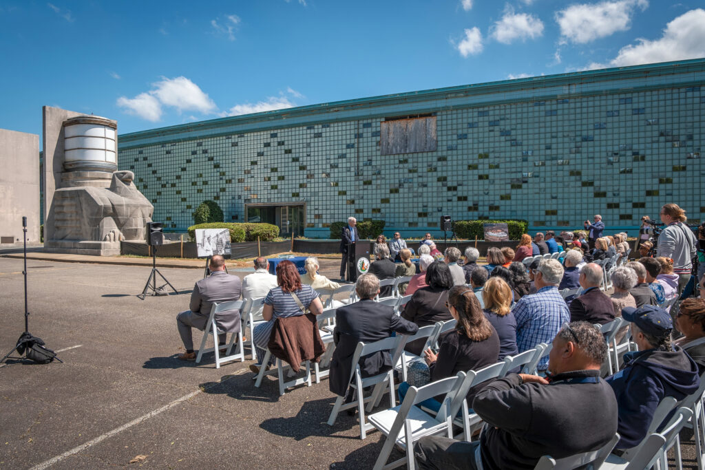 A wide view of people gathered to listen to a speaker beside a building.