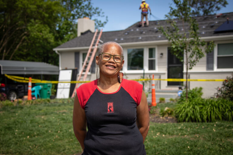 A woman stands in front her her home while solar panels are installed.