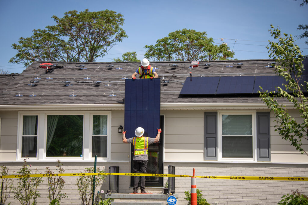 A construction worker passes a solar panel to another construction worker on a roof.