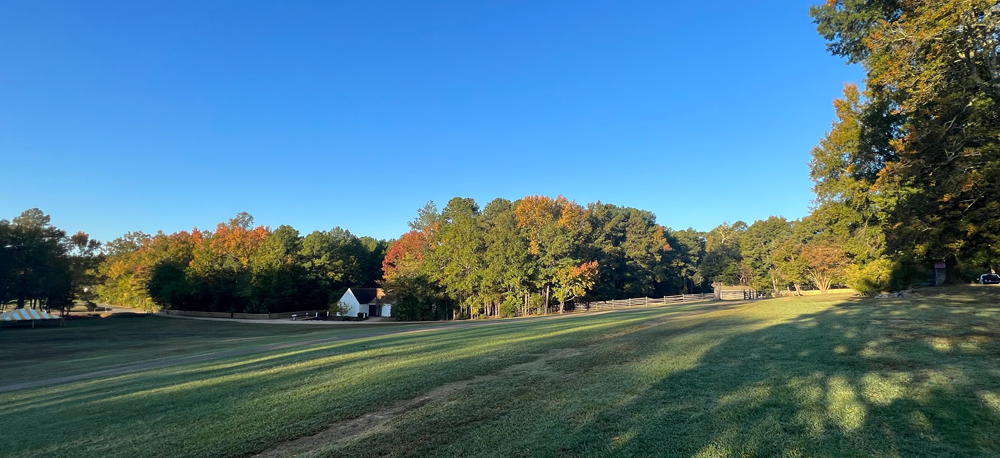 Photo masthead of sweeping green grass with green trees with a few fall color leaves on 3 sides. Nestled in the trees is a white house and  wooden split rail fence.