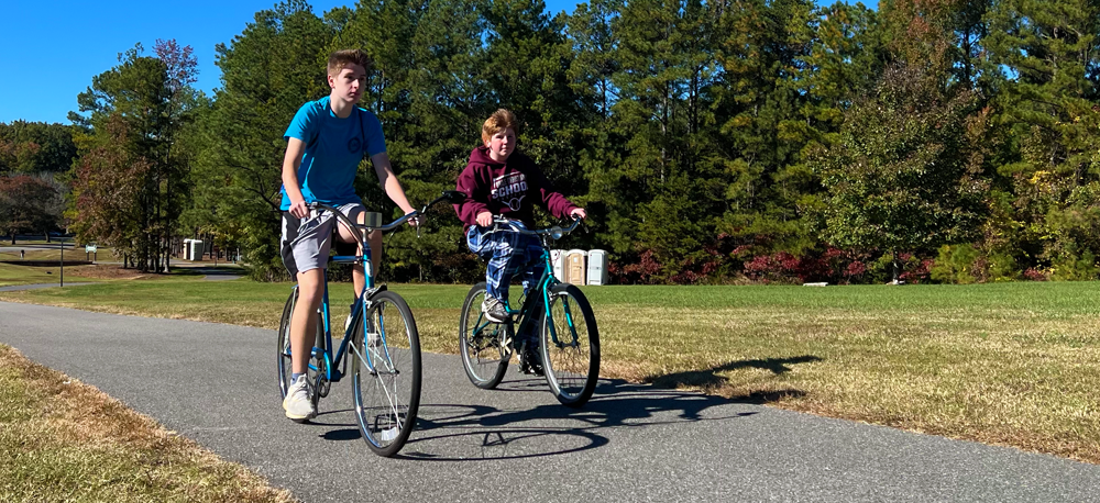 2 adolescent boys each on a green bike riding on a bike pathway next to grass & trees. One boy is wearing a blue shirt & shorts & the other is wearing a maroon hooded sweatshirt with blue plaid pants.