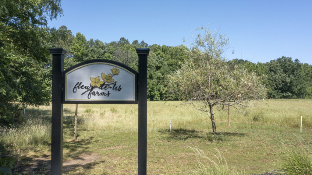 The sign for fleur de lis farms with yellow flowers on the sign and grass and trees in the background.