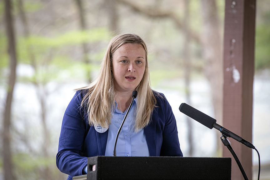 Photo of Kristin Owen, blonde hair, young & wearing a blue jacket, outside at a lectern with a microphone. 