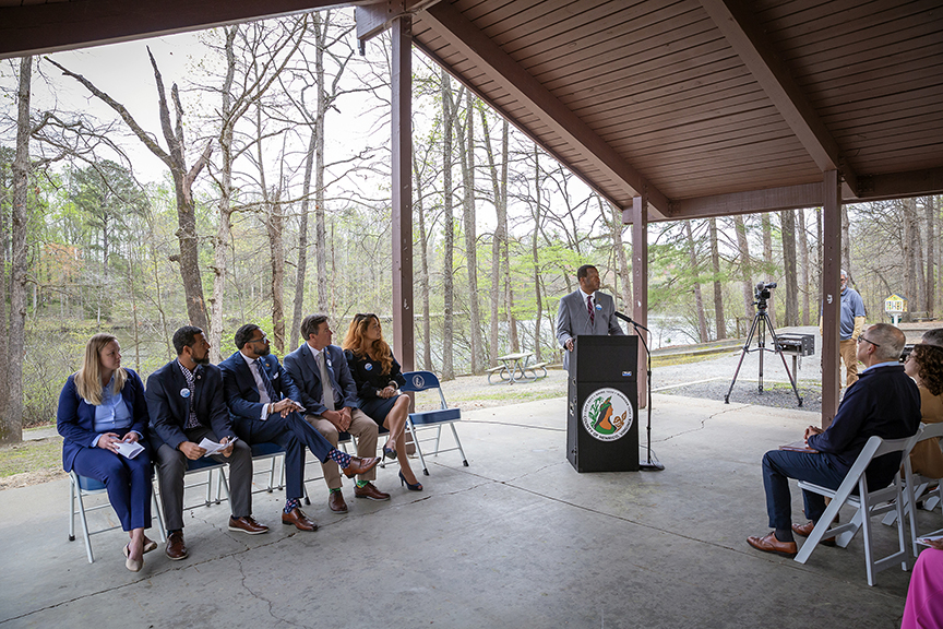 Press conference being held outside under a wooden structure, with 1 person at lectern & ppppeople sitting in chairs