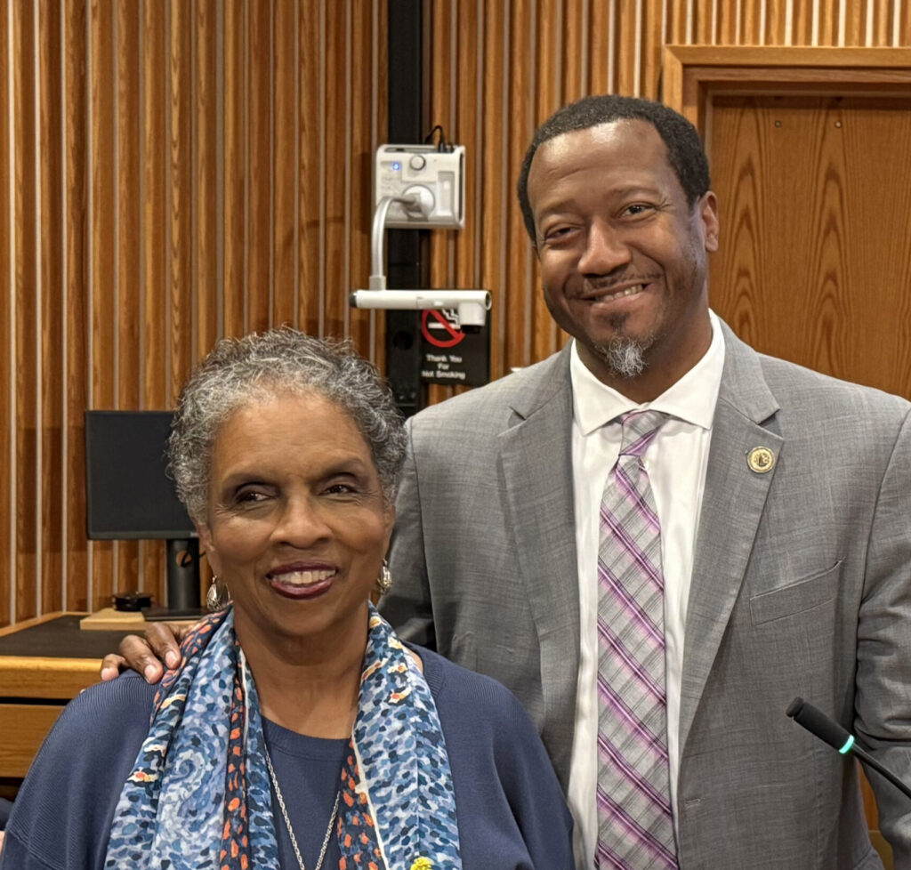 A woman holding flowers and a man smile for a photo in a meeting room.