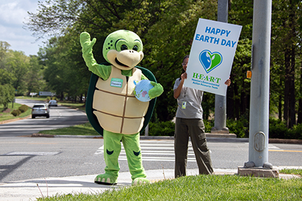 Turtle mascot waves to cars. Person next to him holds Happy Earth Day sign.