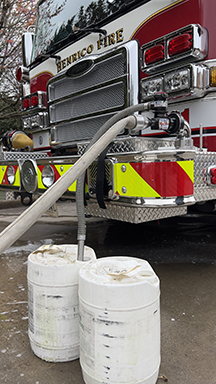 Front of red firetruck with 2 hoses hooked up. 1 hose goes into a white canister with a 2nd canister nearby.