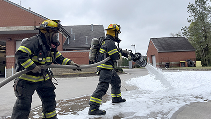 Side photo of 2 firefighters, in full gear, spraying special fire foam, outside, for practice