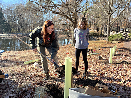 A woman leaning over shoveling dirt next to a yellow tube with a sapling inside & a young girl watching