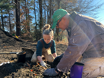 A kneeling man wearing a cap & a coat is planting a sapling with the help of a 
 kneeling young girl