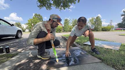 A man wearing a hat kneeling  & a young girl spray painting a template