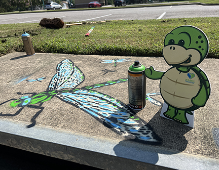 Turtle mascot holding a spray can of paint next to a large dragonfly painted on the top of a storm drain.