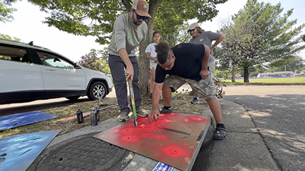 A man wearing a hat  & a teenage boy spray painting a template